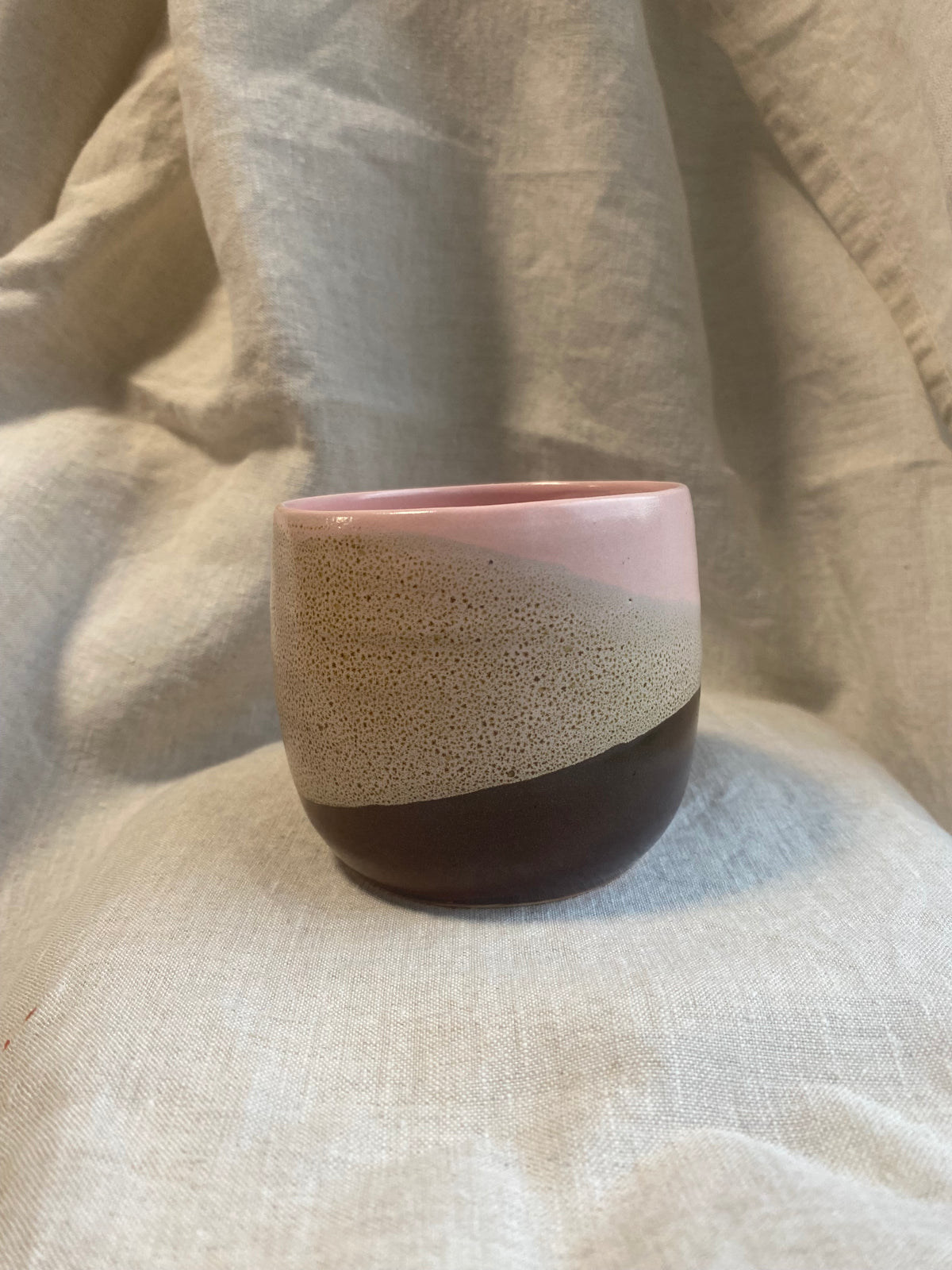 A handmade ceramic cup with a three-color glaze design, featuring a speckled beige middle section, a light pink top section, and a dark brown base with a dimple for holding